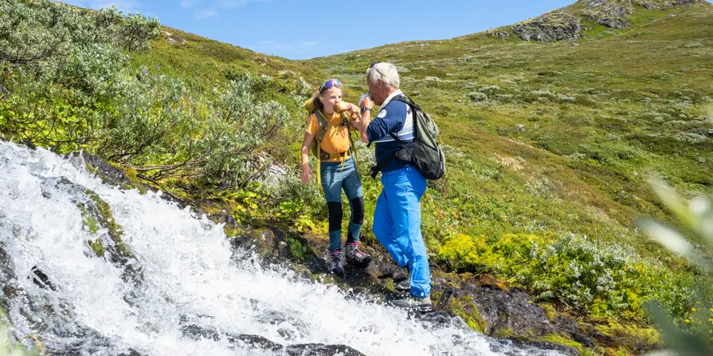 Bestefar og barn tar en drikkepause med fjellvann fra bekken under fjelltur