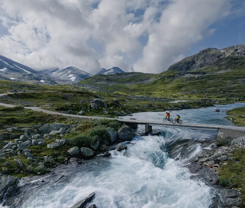 Ungt par på sykkeltur i Koldedalen sykler over bro med storslått bakgtunn