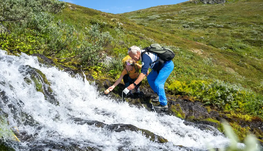 Bedstefar og børn tager en pause for at drikke bjergvand fra bækken under en bjergvandring